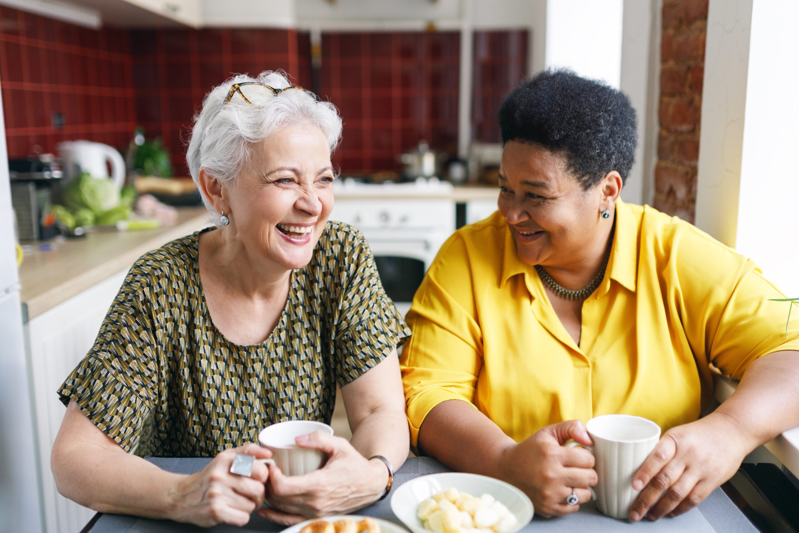 Two senior women laughing together over coffee in a kitchen